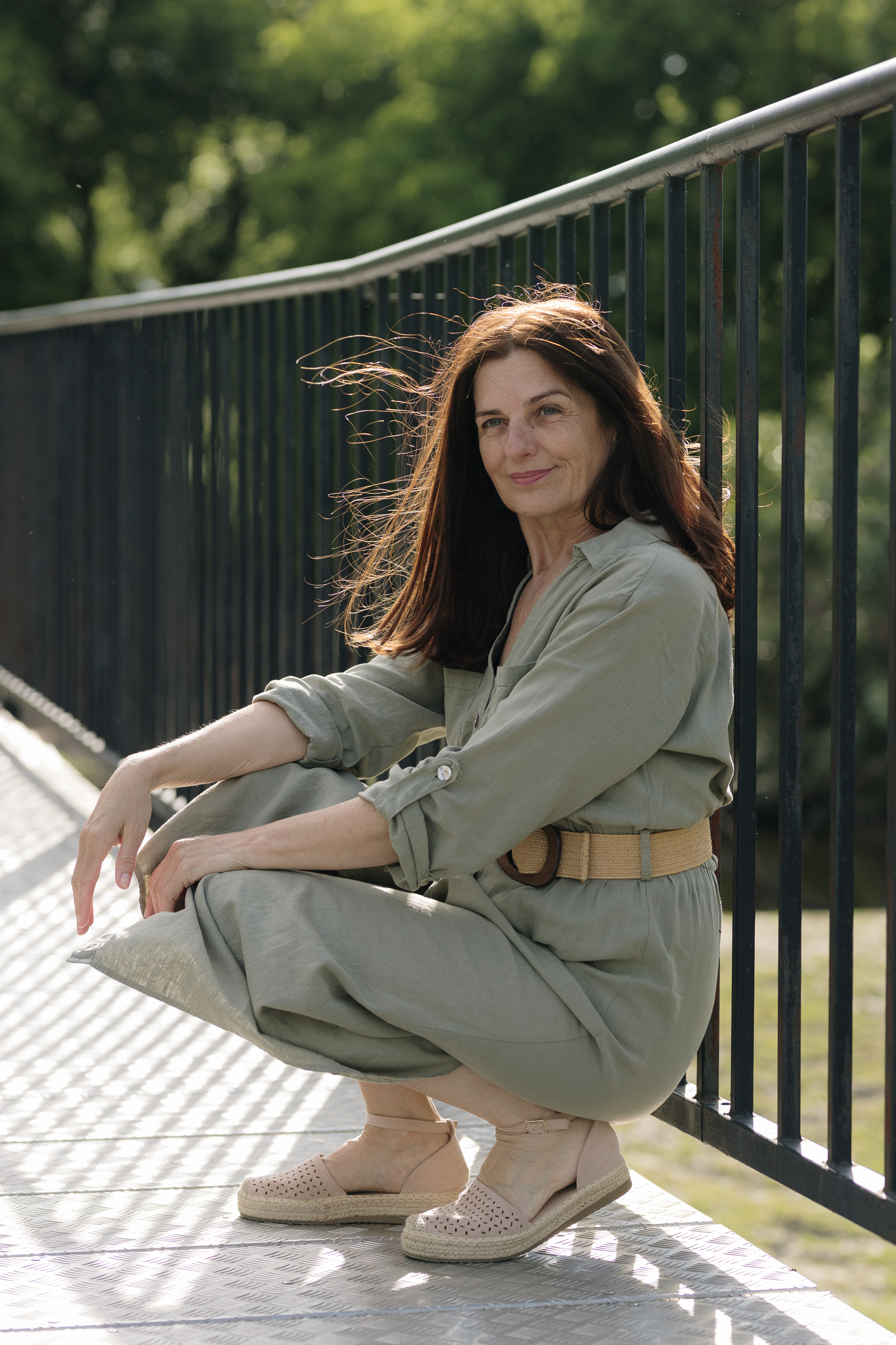 Daniela Cukrov | A woman with long brown hair, wearing a light green dress and beige wedge shoes, squats on a sunlit walkway beside a black railing and green trees, embodying confidence inspired by coaching za žene 45+ in a safe outdoor space.