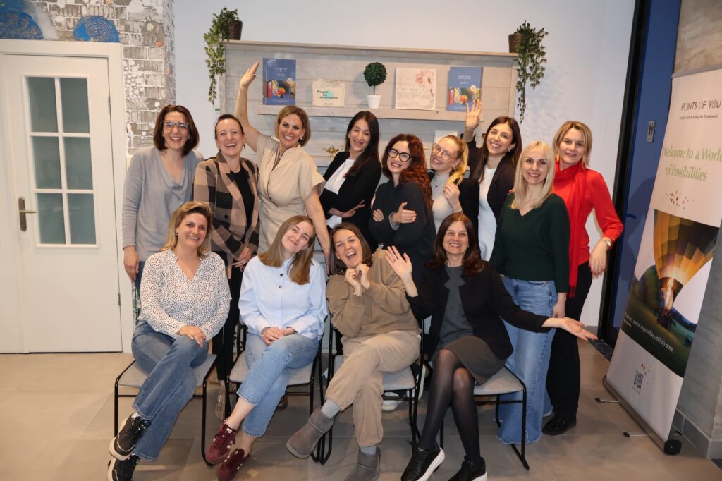 Daniela Cukrov | Thirteen women pose together indoors, smiling and making joyful gestures. Their gathering radiates životna radost i osobni rast žena, with books and plants on a shelf in the background and a colorful poster on the right.
