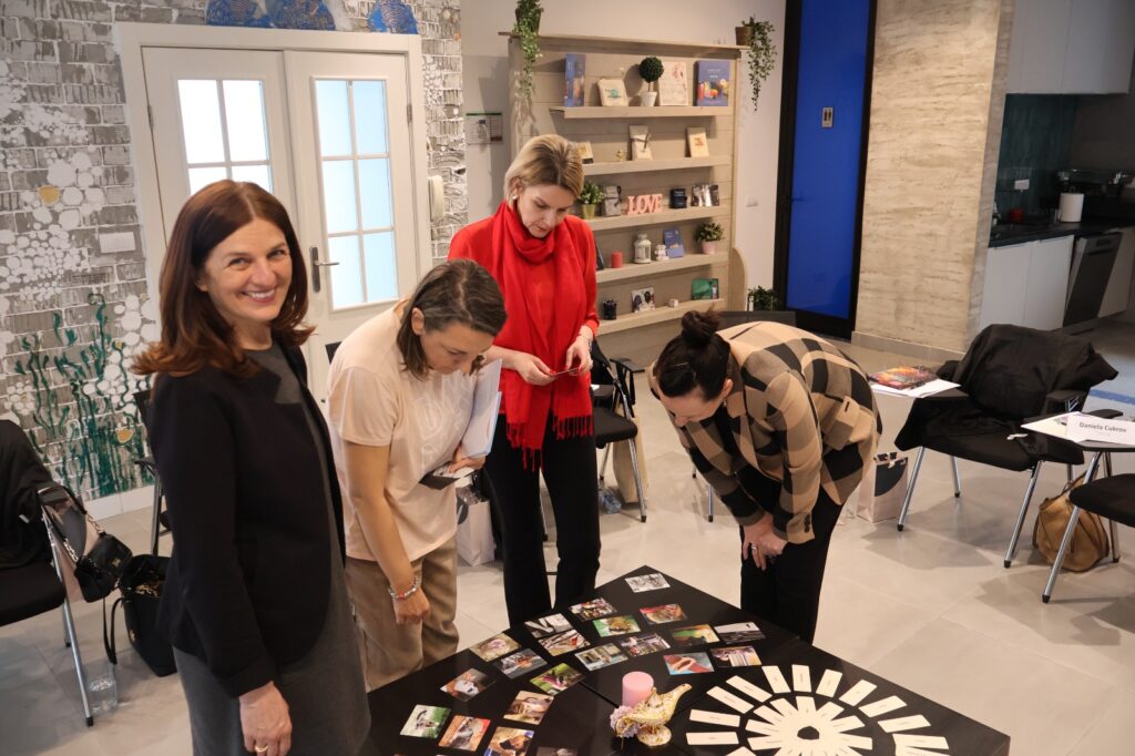 Daniela Cukrov | Four women stand around a black table with assorted cards and objects, examining them closely. Their gathering radiates životna radost i osobni rast žena, as they connect with themselves and each other in a warm, inviting space.