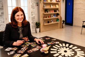 Daniela Cukrov | A smiling woman with red hair sits at a table covered with photographs and cards, holding a photo. The bright room, filled with shelves, plants, and greeting cards, reflects the warmth of radionica osobnog razvoja za žene or coaching za žene 45+.